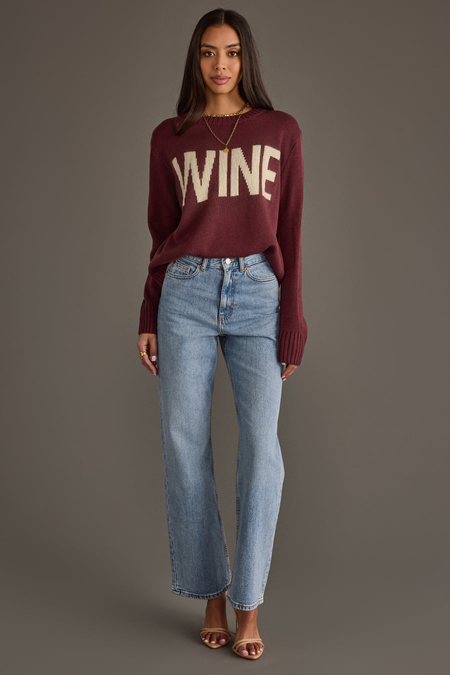 A woman stands against a gray background wearing the Panache Apparel Collection Burgundy & Ivory Wine Sweater, light blue jeans, brown heeled sandals, and a gold necklace.