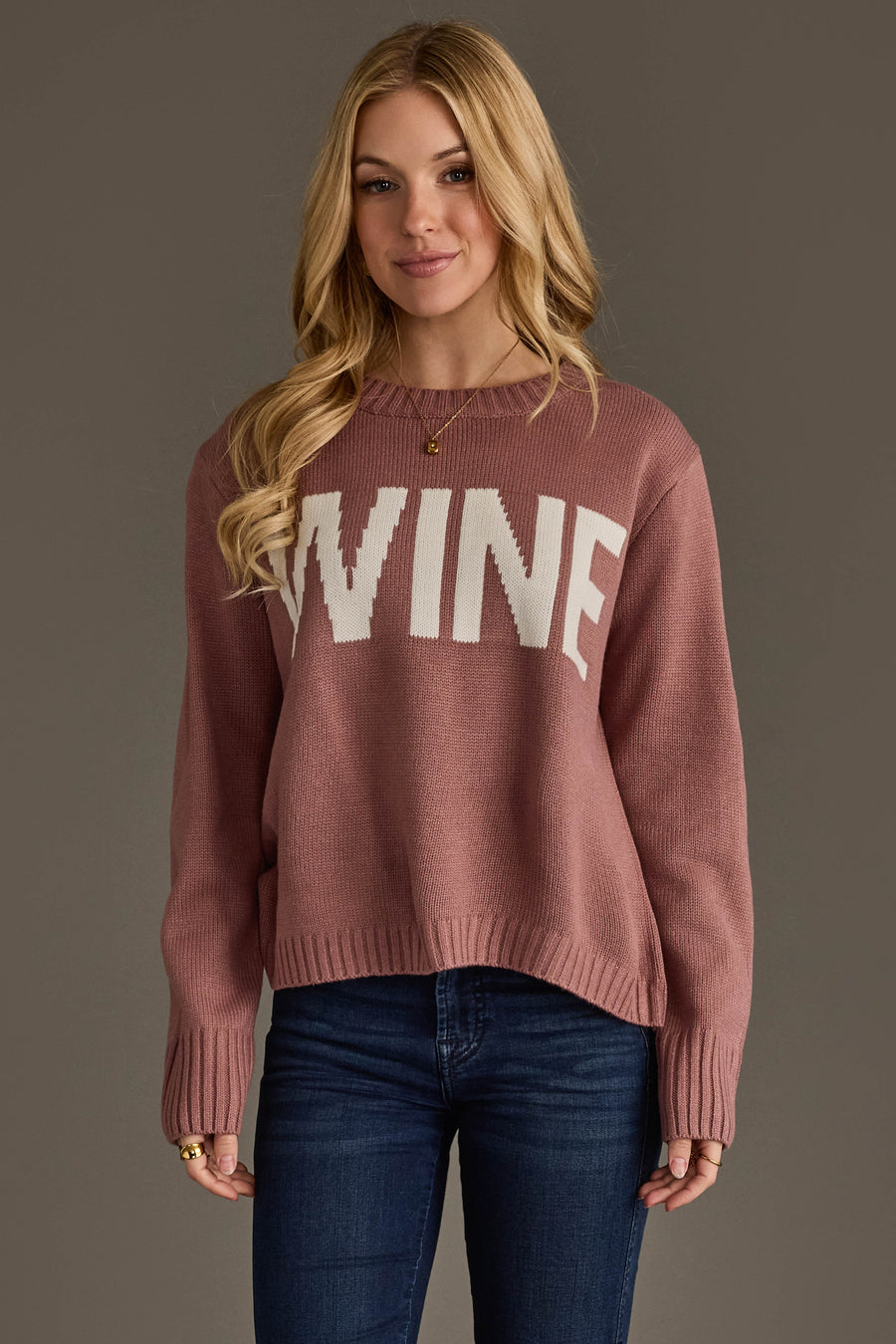 A woman with long blonde hair is wearing the Mauve & White Wine Sweater from Panache Apparel Collection, featuring the word WINE in bold white letters. She pairs it with dark blue jeans and stands against a plain gray background.