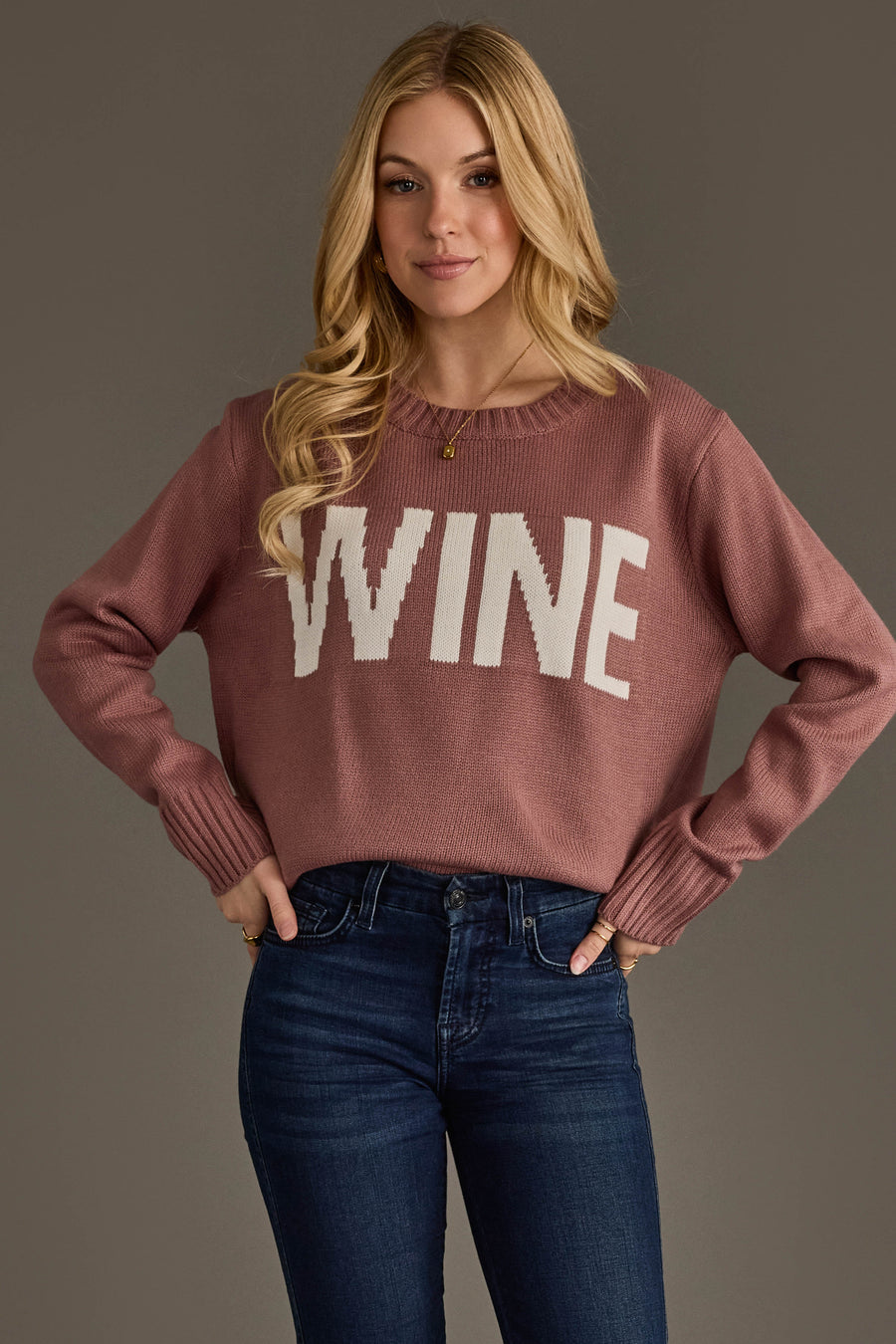 A woman with long blonde hair wears the Panache Apparel Collection Mauve & White Wine Sweater paired with blue jeans, standing with her hands on her hips against a plain gray background.