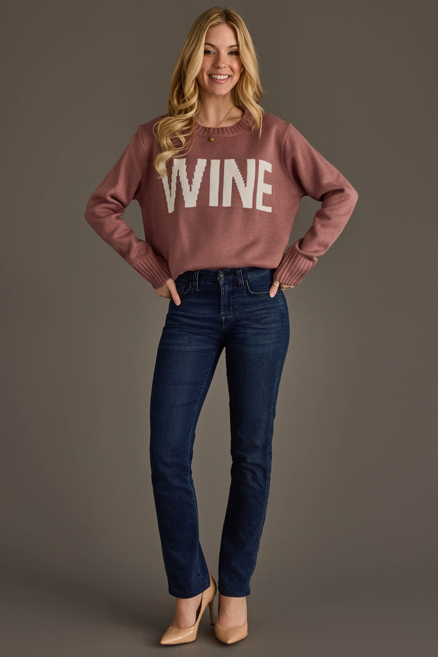 A woman stands smiling, wearing the Mauve & White Wine Sweater from Panache Apparel Collection, paired with dark blue jeans and tan heels. She has long blonde hair and poses with her hands on her hips against a plain background.