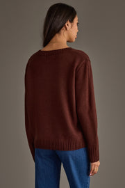 A woman with long dark hair and large hoop earrings stands with her back to the camera, wearing the Panache Apparel Collection’s Coffee Is Calling Sweater in dark brown paired with blue jeans, against a plain gray background.