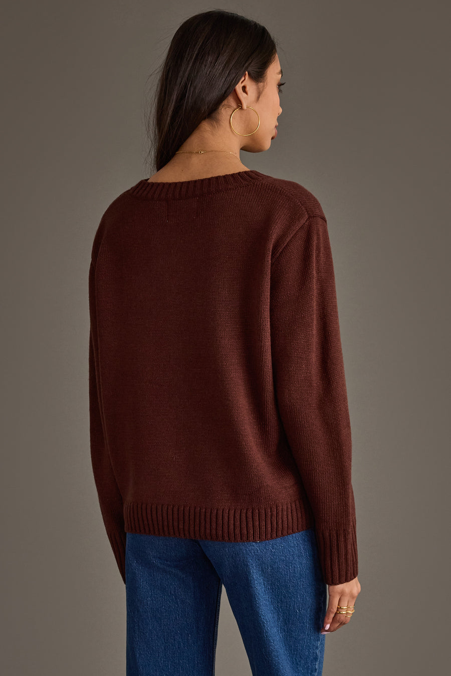 A woman with long dark hair and large hoop earrings stands with her back to the camera, wearing the Panache Apparel Collection’s Coffee Is Calling Sweater in dark brown paired with blue jeans, against a plain gray background.