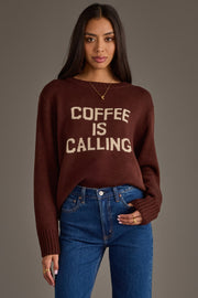 A woman with long dark hair is wearing the Coffee Is Calling Sweater from Panache Apparel Collection a brown sweater featuring the words "COFFEE IS CALLING" in beige letters paired with blue jeans and gold jewelry, standing against a plain gray background.