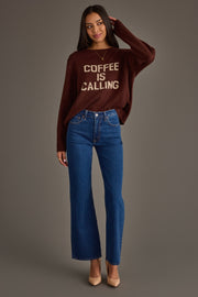 A woman stands smiling against a plain gray background, wearing Panache Apparel Collection’s Coffee Is Calling Sweater in brown, styled with blue high-waisted jeans and beige heels.