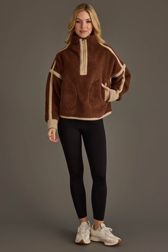 A person wearing the Panache Apparel Collection Brown & Tan Half Zip Sherpa Fleece Sweatshirt, paired with black leggings and white sneakers, stands against a plain gray background.