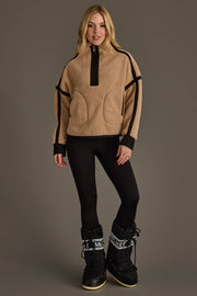 A woman with long blonde hair is smiling while standing against a gray background, wearing the Panache Apparel Collection Tan & Black Half Zip Sherpa Fleece Sweatshirt paired with black leggings and tall black snow boots.