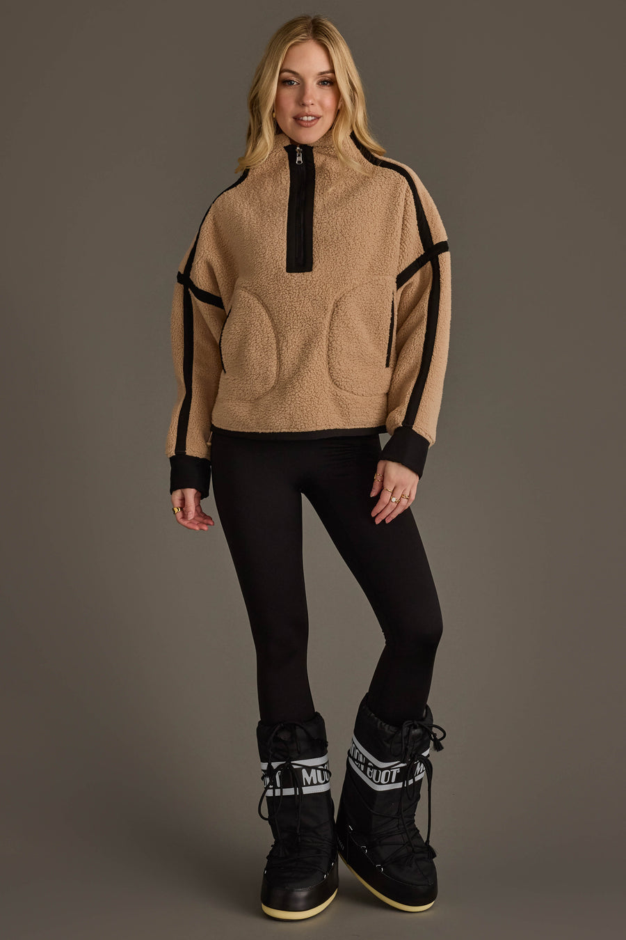 A woman with long blonde hair is smiling while standing against a gray background, wearing the Panache Apparel Collection Tan & Black Half Zip Sherpa Fleece Sweatshirt paired with black leggings and tall black snow boots.