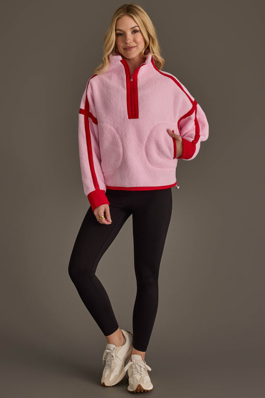 A woman stands smiling against a gray background, wearing the Panache Apparel Collection Light Pink & Red Half Zip Sherpa Fleece Sweatshirt, paired with black leggings and beige sneakers.
