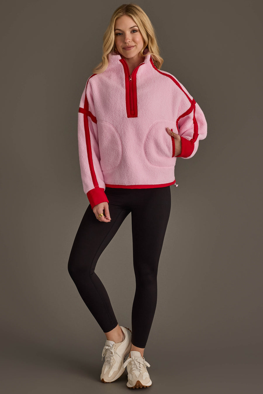 A woman stands smiling against a gray background, wearing the Panache Apparel Collection Light Pink & Red Half Zip Sherpa Fleece Sweatshirt, paired with black leggings and beige sneakers.