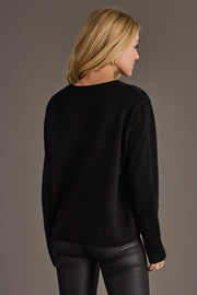 A woman with long blonde hair, wearing the Panache Apparel Collection’s Spooky Sweater and black leather pants, stands facing away from the camera against a plain gray background.