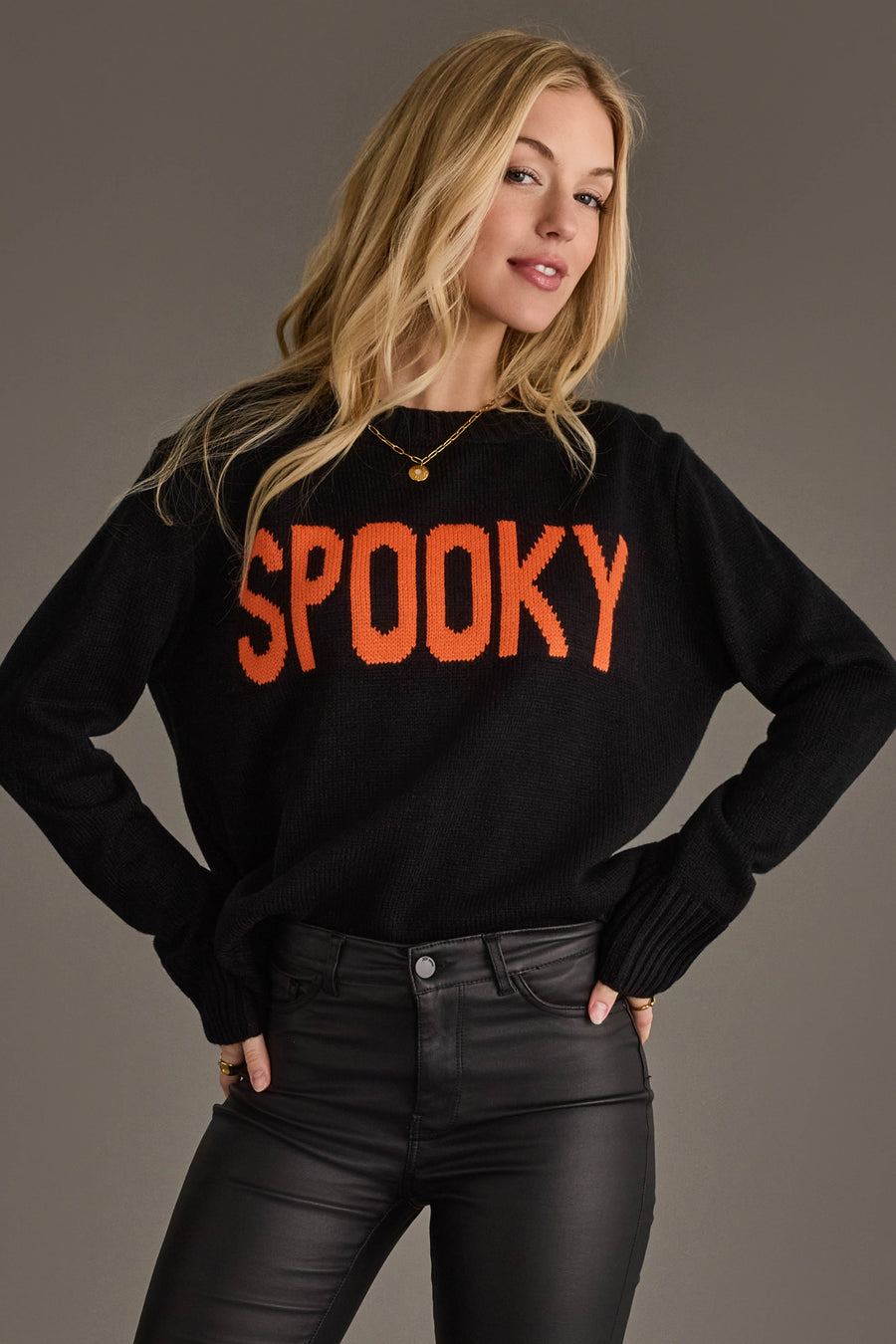 A woman models the Panache Apparel Collection’s Spooky Sweater, a black knit featuring bold orange “SPOOKY” lettering, paired with black pants, a gold necklace, and rings, as she poses with one hand on her hip against a plain gray background.