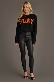 A woman stands against a gray background wearing the Panache Apparel Collection’s Spooky Sweater, a black sweater featuring “SPOOKY” in bold orange letters, paired with black leather pants and black open-toe heels.