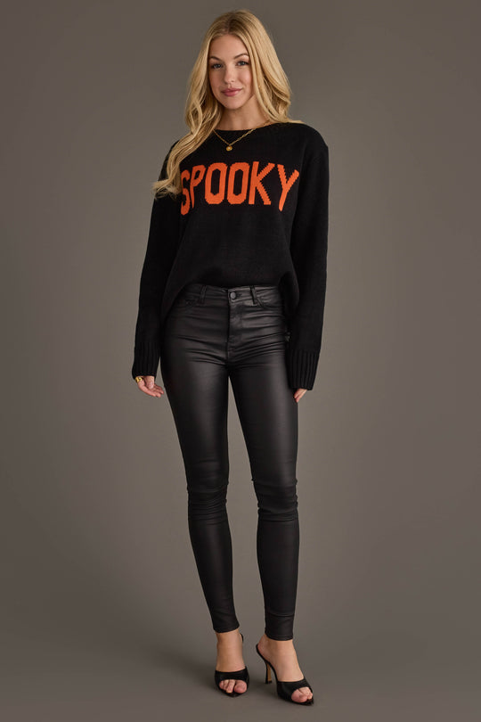 A woman stands against a gray background wearing the Panache Apparel Collection’s Spooky Sweater, a black sweater featuring “SPOOKY” in bold orange letters, paired with black leather pants and black open-toe heels.