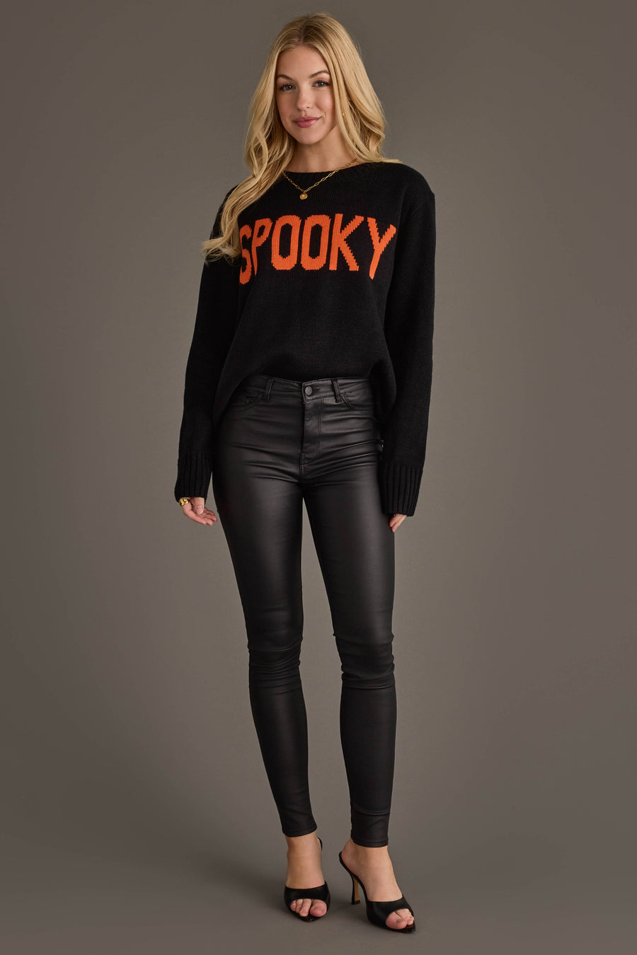 A woman stands against a gray background wearing the Panache Apparel Collection’s Spooky Sweater, a black sweater featuring “SPOOKY” in bold orange letters, paired with black leather pants and black open-toe heels.