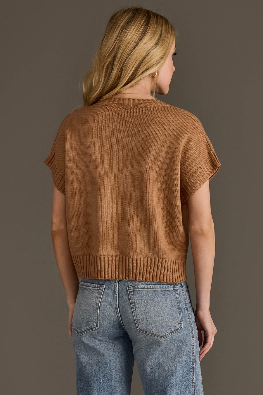 A back view of a blonde woman wearing the Panache Apparel Collection's Coffee Weather Short Sleeve Sweatshirt a camel-colored knit sweater vest featuring a ribbed hem and neckline paired with light-wash blue jeans, set against a solid dark grey background.
