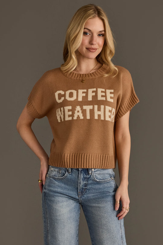 A woman with blonde hair is wearing the Coffee Weather Short Sleeve Sweatshirt by Panache Apparel Collection a brown knit top with "COFFEE WEATHER" printed in white paired with blue jeans and standing against a plain gray background.
