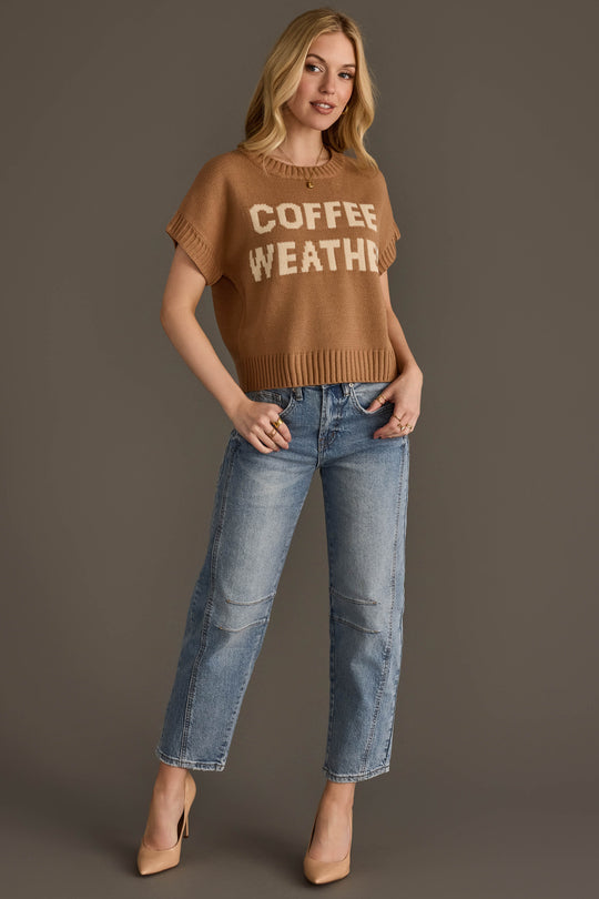 A woman stands against a brown background wearing the Panache Apparel Collection's Coffee Weather Short Sleeve Sweatshirt, paired with blue jeans and tan heels. She has long blonde hair and is smiling slightly with one hand in her pocket.