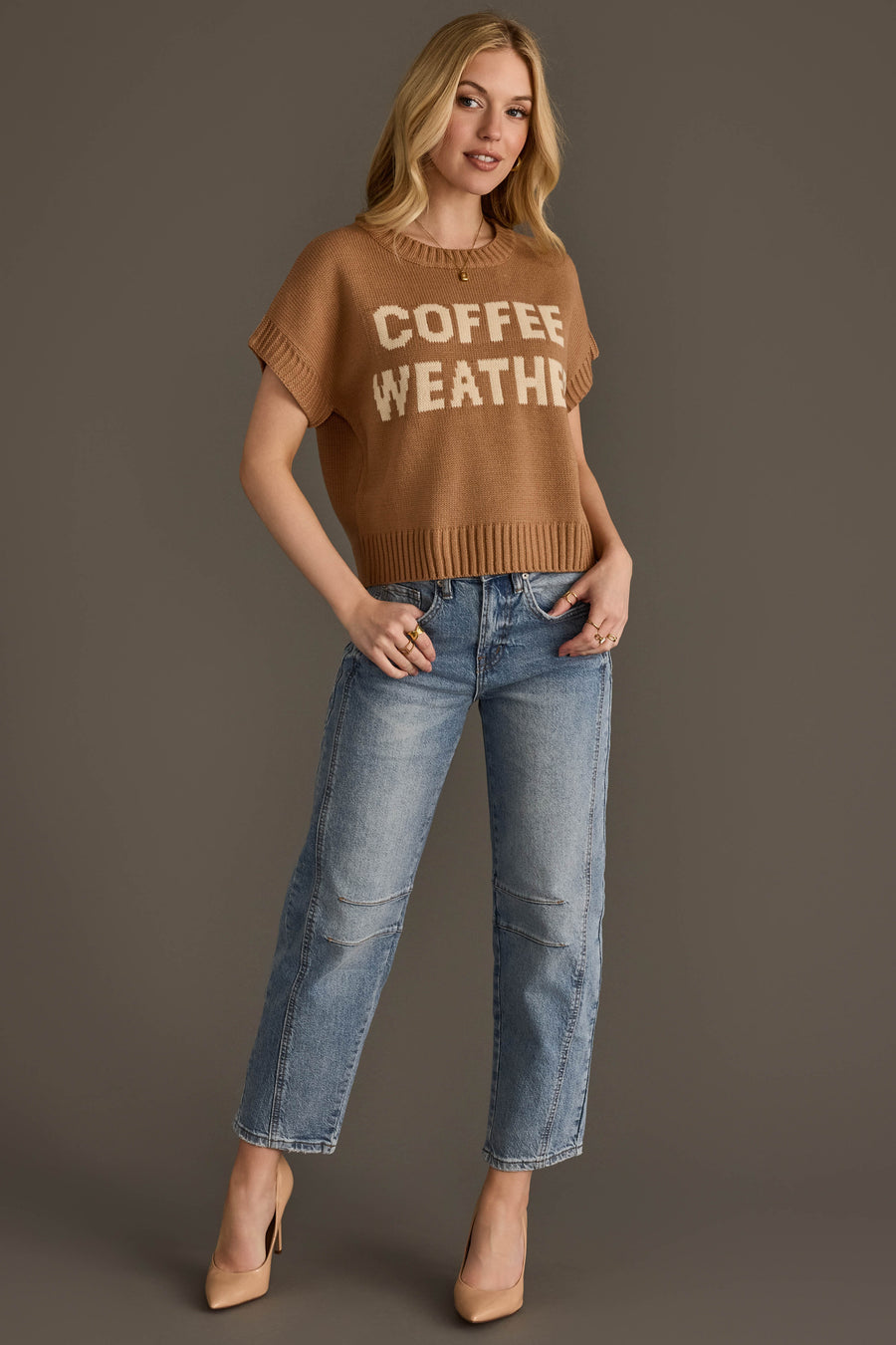 A woman stands against a brown background wearing the Panache Apparel Collection's Coffee Weather Short Sleeve Sweatshirt, paired with blue jeans and tan heels. She has long blonde hair and is smiling slightly with one hand in her pocket.