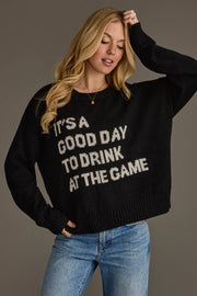 A woman models the Panache Apparel Collection's "Drink At The Game" Lightweight Sweater—a black top featuring the phrase "ITS A GOOD DAY TO DRINK AT THE GAME" in white lettering—styled with light blue jeans against a plain background.