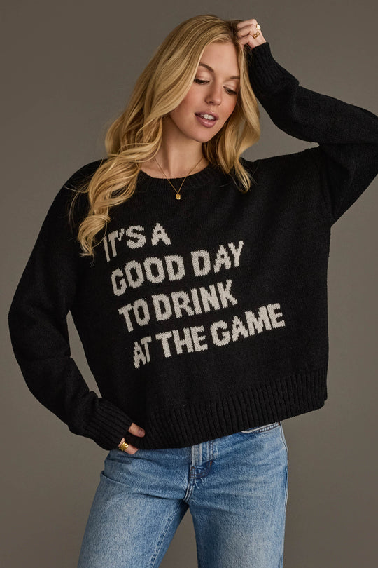 A woman models the Panache Apparel Collection's "Drink At The Game" Lightweight Sweater—a black top featuring the phrase "ITS A GOOD DAY TO DRINK AT THE GAME" in white lettering—styled with light blue jeans against a plain background.