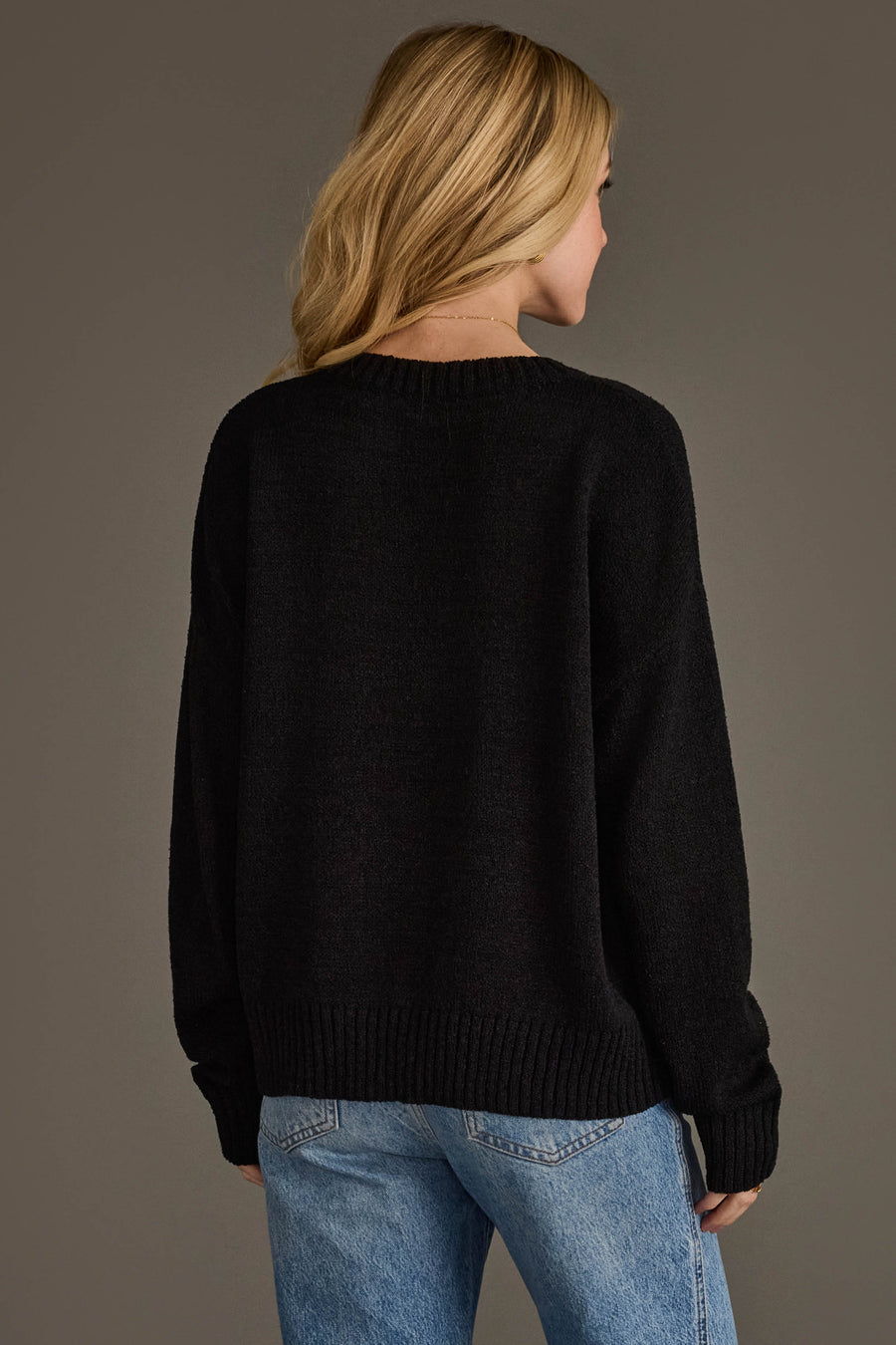 A person with long blonde hair, wearing the Panache Apparel Collection's Drink At The Game Lightweight Sweater in black and blue jeans, stands facing away from the camera against a plain background.