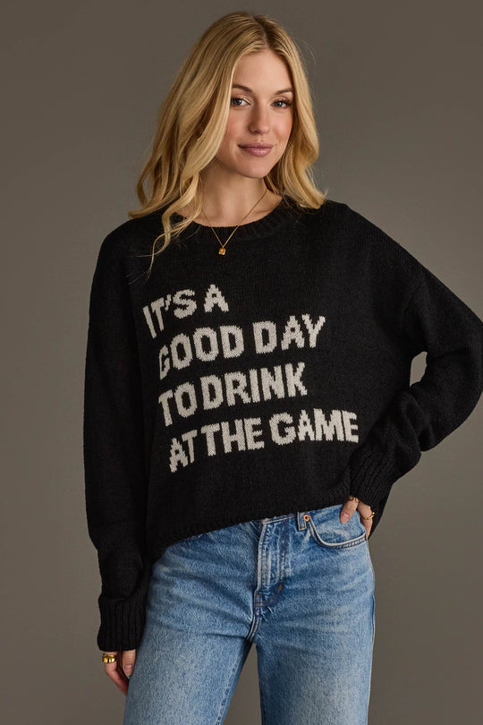 A woman smiles against a plain background with one hand in her jeans pocket, wearing the Panache Apparel Collection's Drink At The Game Lightweight Sweater in black, featuring the phrase "ITS A GOOD DAY TO DRINK AT THE GAME.