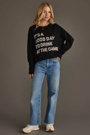 A woman wearing the Panache Apparel Collection’s "Drink At The Game" Lightweight Sweater, paired with light blue jeans and white sneakers, stands against a plain gray background, smiling with one hand in her pocket.