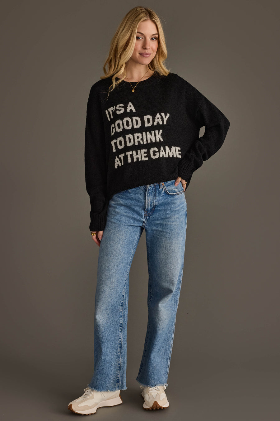 A woman wearing the Panache Apparel Collection’s "Drink At The Game" Lightweight Sweater, paired with light blue jeans and white sneakers, stands against a plain gray background, smiling with one hand in her pocket.