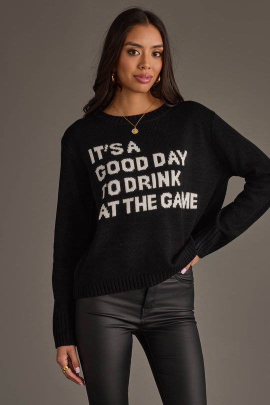 Model wearing the “Drink At The Game Sweater” by Panache Apparel Collection—a black sweater with white "ITS A GOOD DAY TO DRINK AT THE GAME" text—paired with black leather pants, posed against a plain gray background.