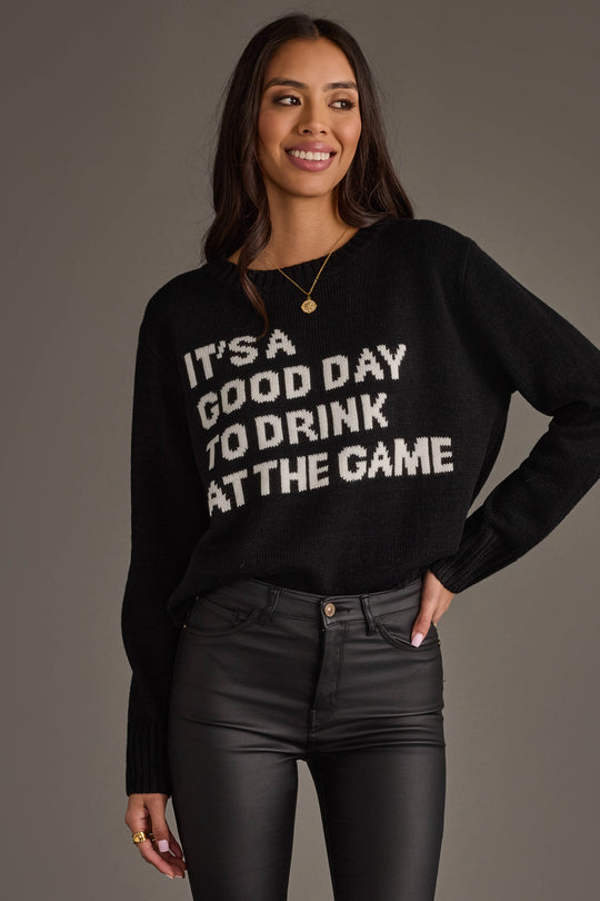 A woman with long dark hair smiles while wearing the Panache Apparel Collection's "Drink At The Game Sweater," a black sweater featuring white text that says, "ITS A GOOD DAY TO DRINK AT THE GAME." She is also dressed in black pants and stands against a plain background.