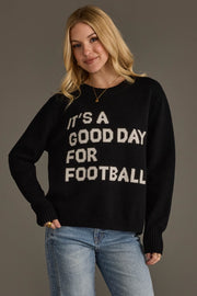 A woman with long blonde hair is wearing the Panache Apparel Collection's "It's A Good Day For Football" soft sweater in black, featuring white lettering, paired with blue jeans and standing against a plain gray background.