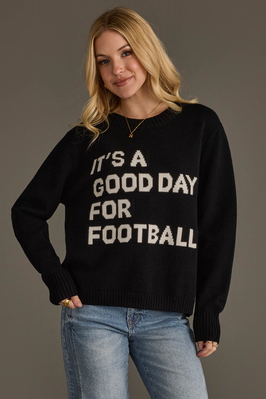 A woman with long blonde hair is wearing the Panache Apparel Collection's "It's A Good Day For Football" soft sweater in black, featuring white lettering, paired with blue jeans and standing against a plain gray background.