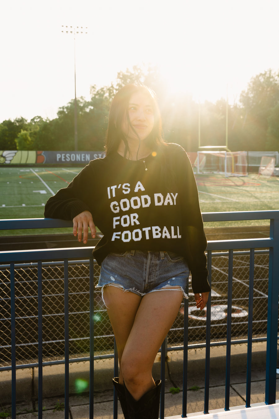 Wearing the Panache Apparel Collection's "It's A Good Day For Football" Soft Sweater, a woman stands by a fence at the football field, sunlight glowing behind her.