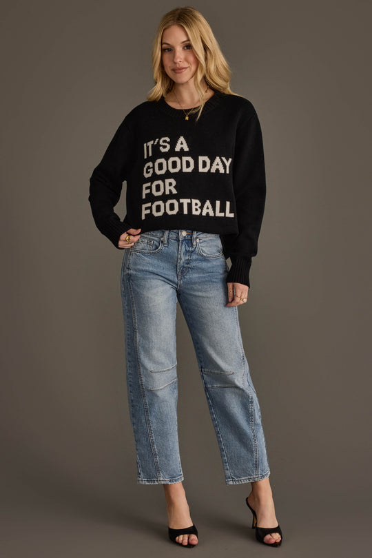 A woman stands smiling against a plain gray background, wearing the Panache Apparel Collection "It's A Good Day For Football" soft sweater in black, paired with light blue jeans and black heels.