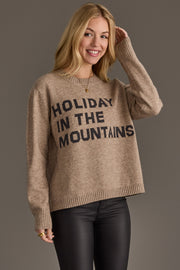 A woman wearing a Taupe Holiday In The Mountains Soft Sweater featuring the text HOLIDAY IN THE MOUNTAINS in black letters.