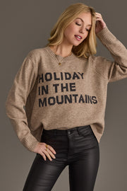 A person wearing a Taupe Holiday In The Mountains Soft Sweater, which is a taupe sweater with the text HOLIDAY IN THE MOUNTAINS in black letters.