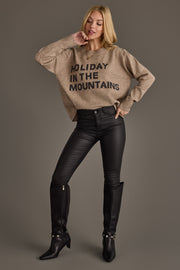 A woman wears a Taupe Holiday In The Mountains Soft Sweater with black text, paired with black skinny pants and black knee-high boots.