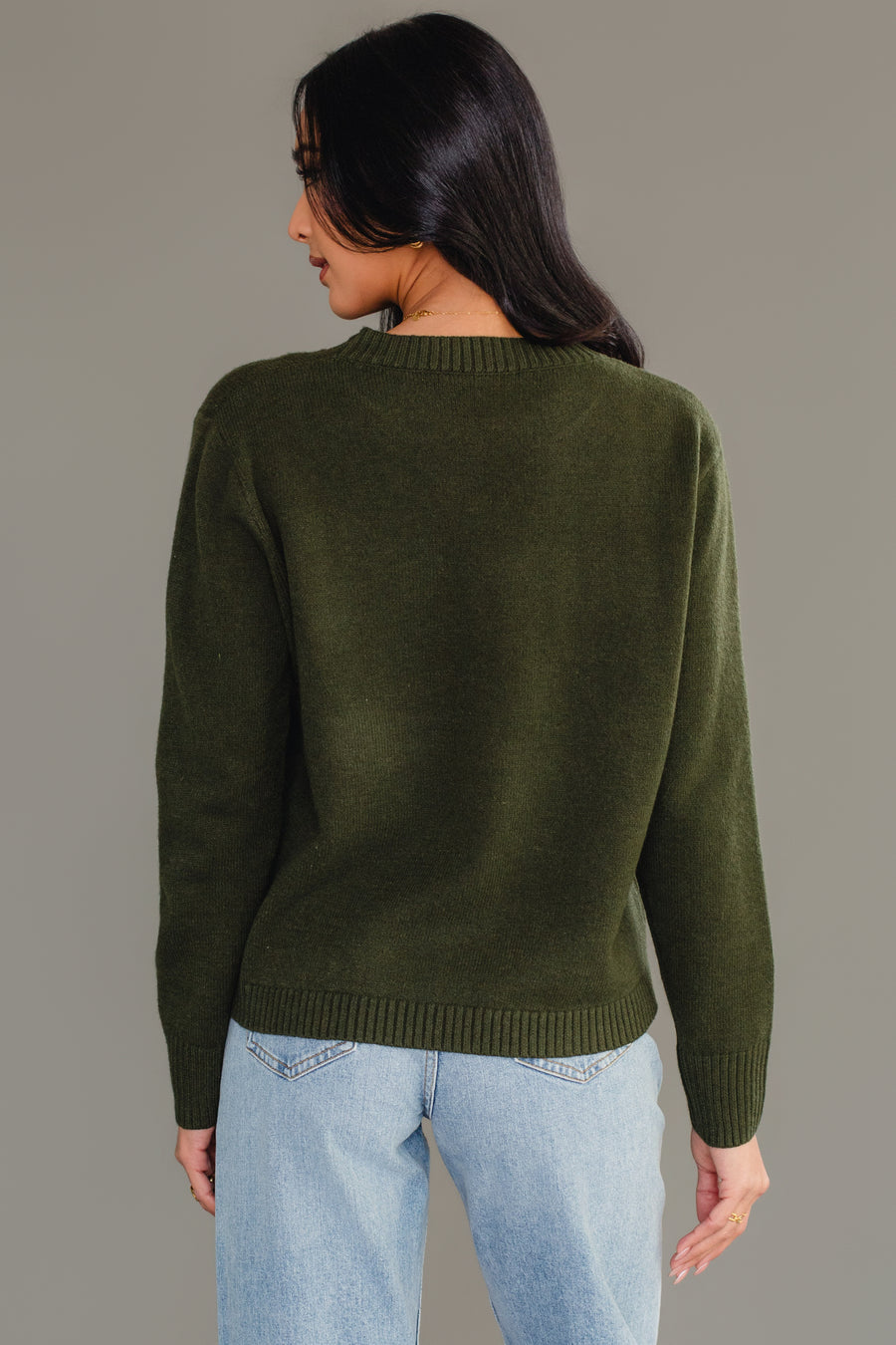 A person wears a Dark Green Holiday In The Mountains Soft Sweater featuring a crew neck and ribbed cuffs, paired with light wash blue jeans.