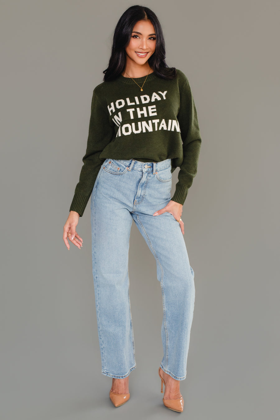 A person wearing a Dark Green Holiday In The Mountains Soft Sweater paired with light-wash blue jeans and tan high heels.
