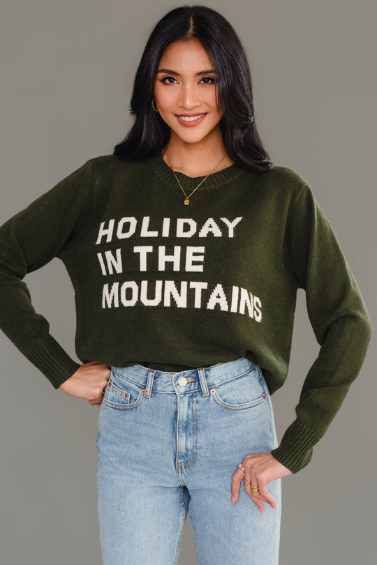 A woman wearing a dark green Dark Green Holiday In The Mountains Soft Sweater with the words HOLIDAY IN THE MOUNTAINS in white text on the chest.