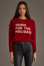 A person wearing a red Home For The Holidays Soft Sweater with white text, paired with black pants.
