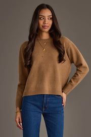 A woman with long brown hair stands against a plain gray background, looking slightly to her right with a soft smile, wearing the Panache Apparel Collection's Tan Soft Sweater paired with blue jeans, gold necklaces, and bracelets.