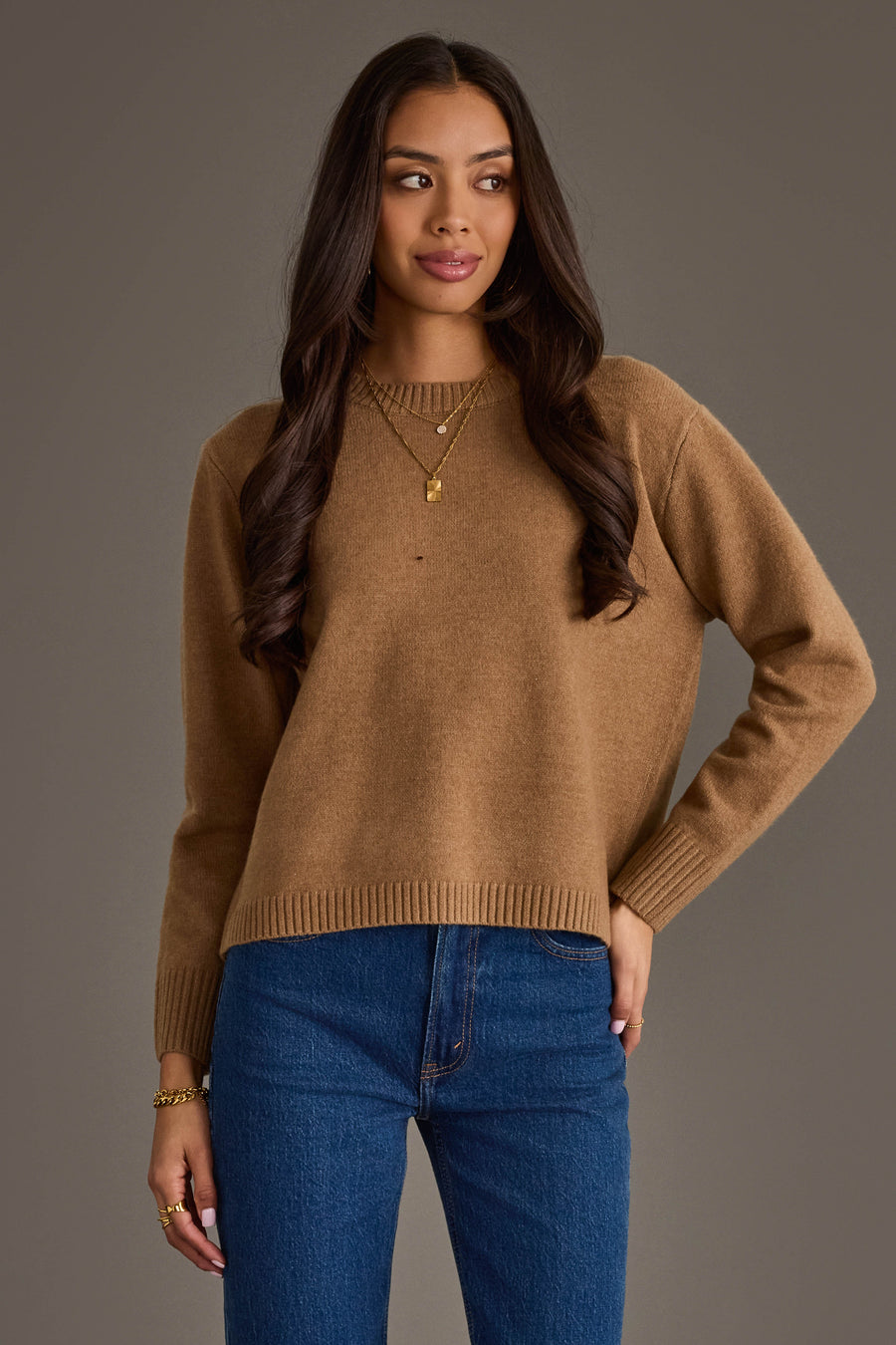 A woman with long brown hair stands against a plain gray background, looking slightly to her right with a soft smile, wearing the Panache Apparel Collection's Tan Soft Sweater paired with blue jeans, gold necklaces, and bracelets.