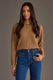 A woman with long brown hair, dressed in the Panache Apparel Collection's Tan Soft Sweater, blue jeans, and gold jewelry, stands against a plain gray background, smiling softly at the camera.