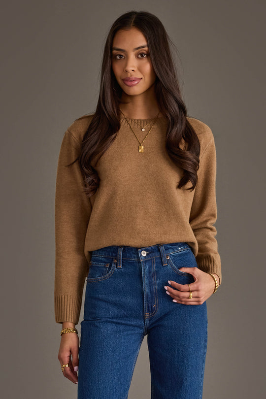 A woman with long brown hair, dressed in the Panache Apparel Collection's Tan Soft Sweater, blue jeans, and gold jewelry, stands against a plain gray background, smiling softly at the camera.
