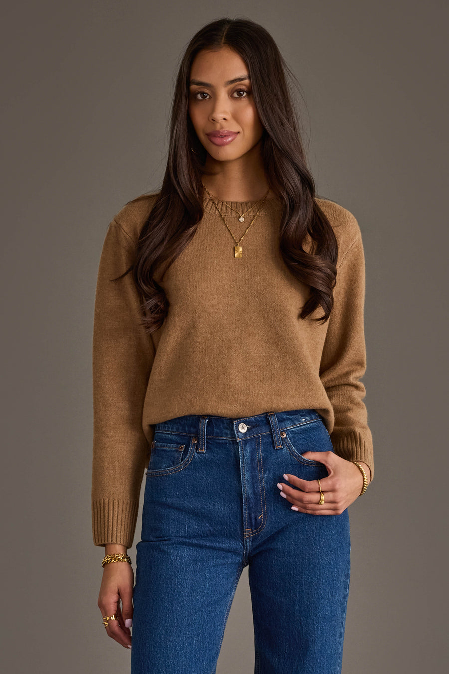 A woman with long brown hair, dressed in the Panache Apparel Collection's Tan Soft Sweater, blue jeans, and gold jewelry, stands against a plain gray background, smiling softly at the camera.