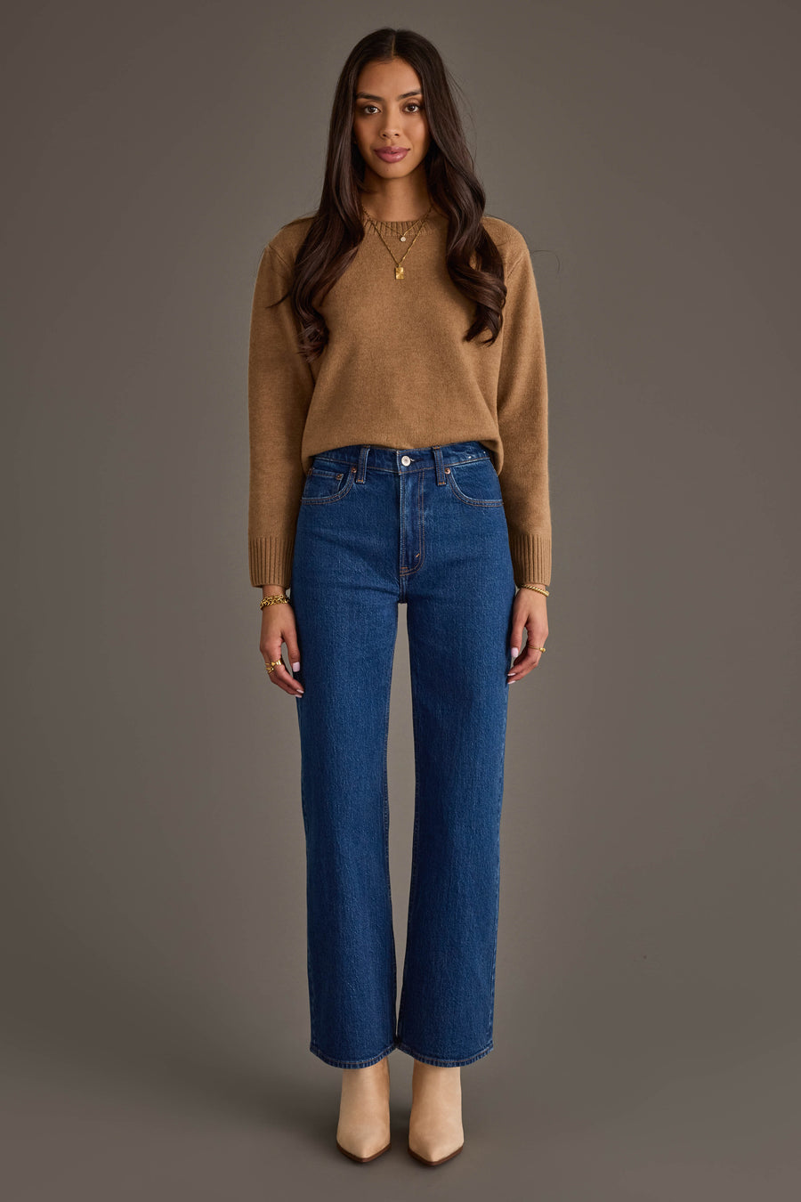 A woman stands facing forward, wearing the Panache Apparel Collection Tan Soft Sweater, paired with blue high-waisted jeans and beige shoes; her long dark hair contrasts with the neutral background.
