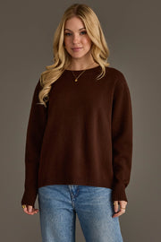 A person with long blonde hair, dressed in the Panache Apparel Collection's Brown Soft Sweater along with blue jeans and gold jewelry, stands in front of a plain gray background, smiling gently at the camera.