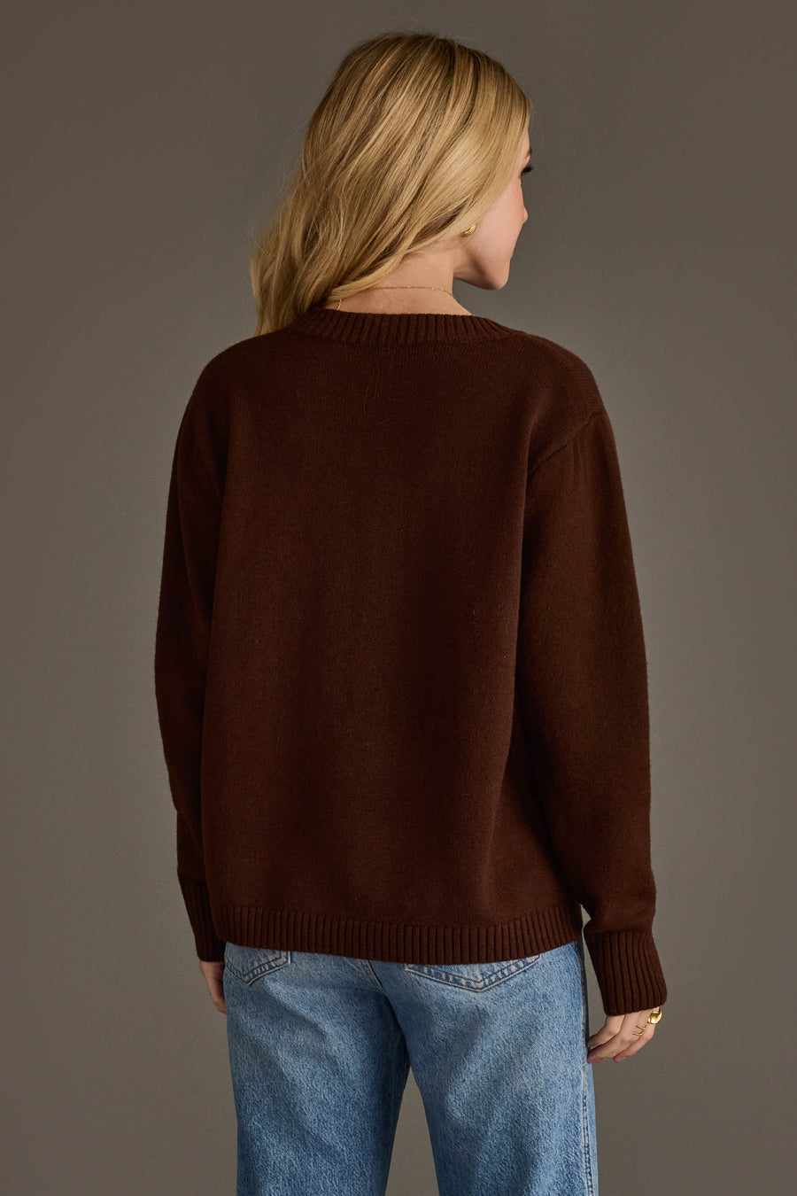 A person is seen from behind, standing against a plain gray background, wearing the Panache Apparel Collection's Brown Soft Sweater paired with light blue jeans.