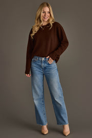 A woman with long blonde hair stands and smiles against a plain gray background, wearing the Panache Apparel Collection's Brown Soft Sweater, paired with blue high-waisted jeans and tan heels.